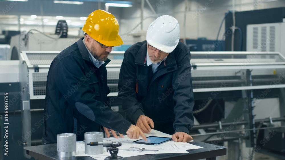 Two engineers discuss a blueprint while checking information on a tablet computer in a factory.