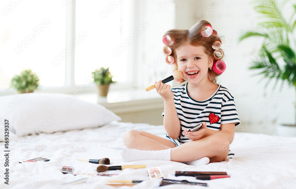 美丽有趣的小女孩戴着卷发器在床上化妆大笑