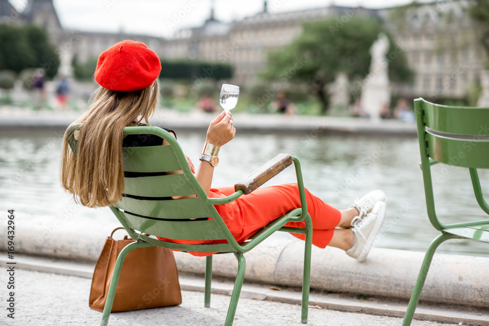 戴着红色帽子，穿着红色裤子，端着一杯葡萄酒坐在土族著名的绿色椅子上的年轻时尚女性