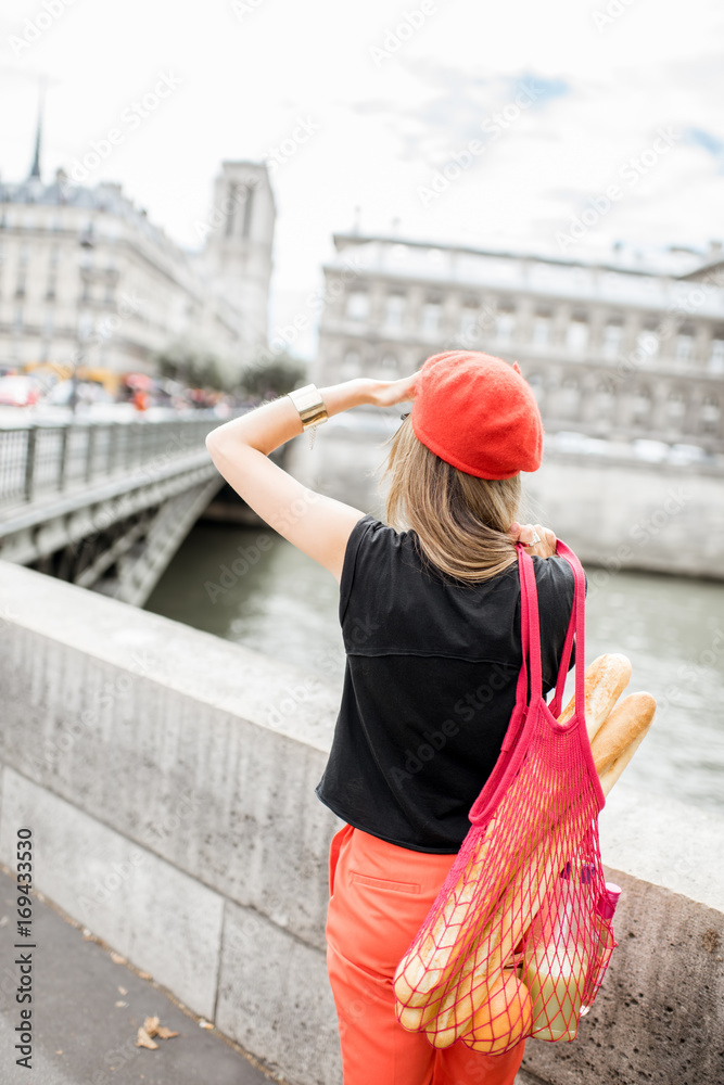 巴黎街头，一位年轻时尚的女士戴着红色帽子，背着装满法国食物的网袋站在后面