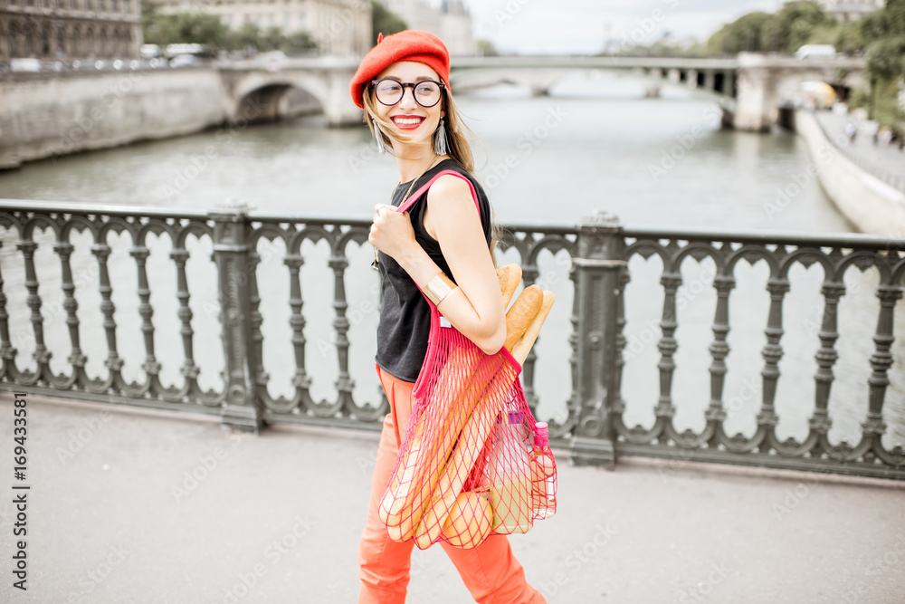巴黎桥上，一位戴着红色帽子的年轻时尚女士，提着装满法国食物的网袋行走