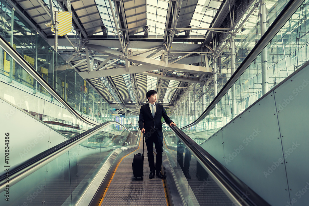 Asian young businessman with luggage down the escalator in airport. Business travel concept.