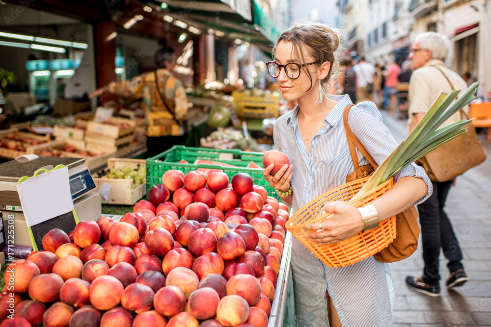 法国菜市场上，一位年轻女士拿着篮子站在那里挑选新鲜的桃子