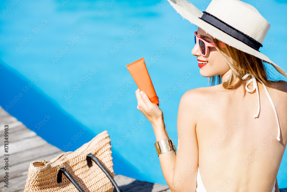 戴着太阳帽的女人拿着防晒乳液坐在蓝色水盆附近。防晒防晒霜