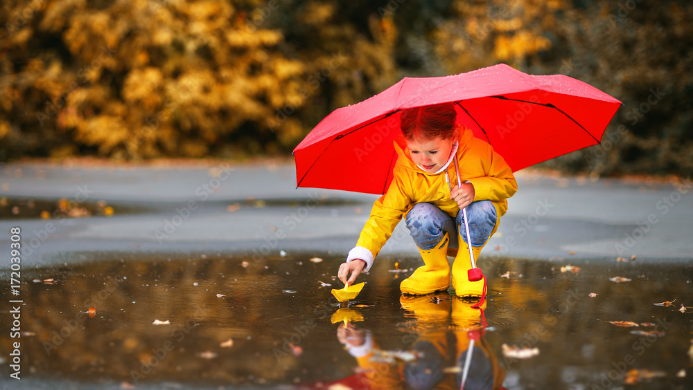 秋天水坑里带着雨伞和纸船的快乐小女孩在大自然中