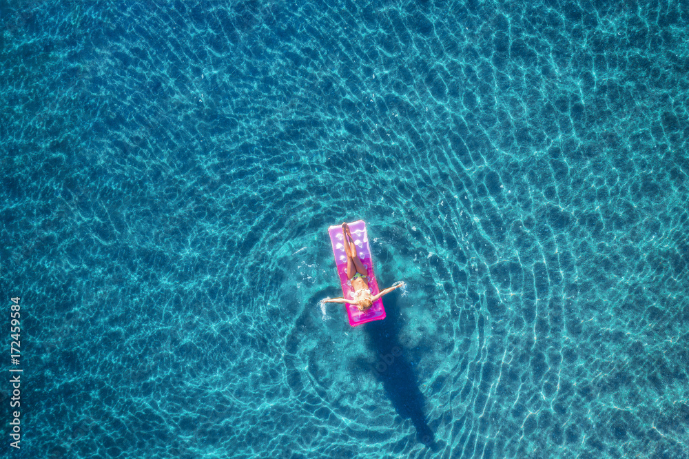 透明绿松石海中，一名年轻女子在粉色充气床垫上游泳的鸟瞰图