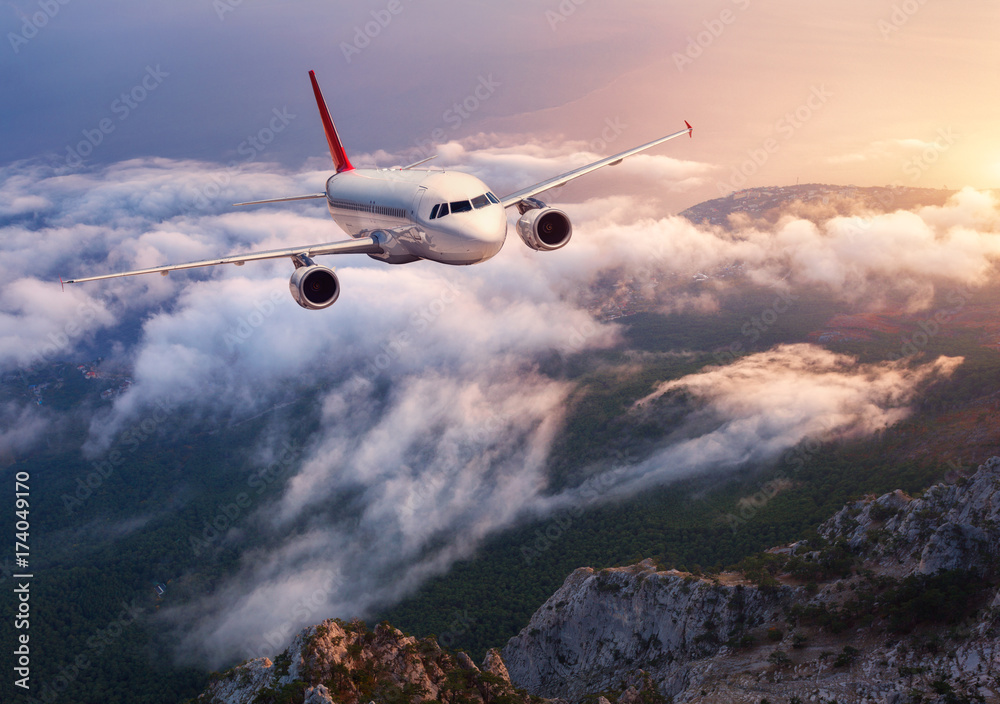 Beautiful airplane is flying over low clouds at sunset. Landscape with passenger airplane, rocks, fo