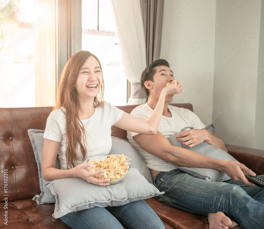 一对亚洲夫妇坐在家里的沙发上看电影，兴高采烈地给男朋友拿爆米花
