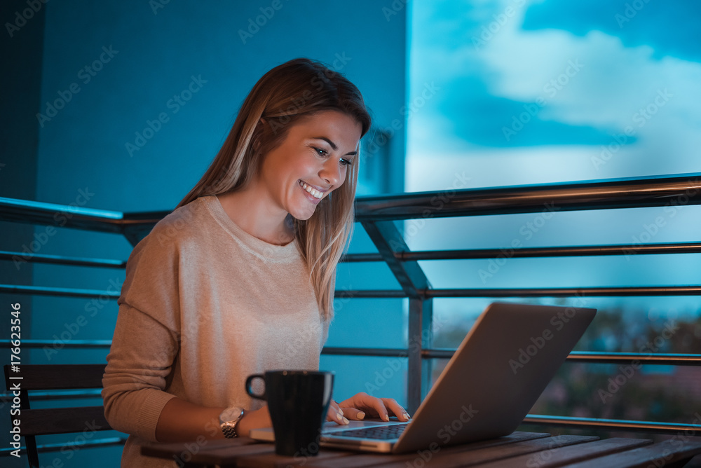 黄昏时分，女人在笔记本电脑上工作，在阳台上微笑。高ISO图像。