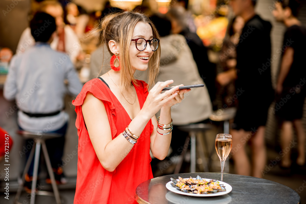 一名身穿红色连衣裙的年轻女子坐在食品市场旁拍摄一盘贻贝和一杯葡萄酒