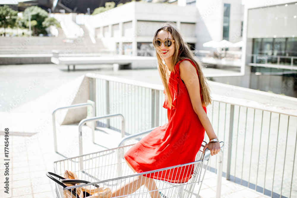 Lifestyle portrait of a young woman in red dress having fun in the shopping cart outdoors