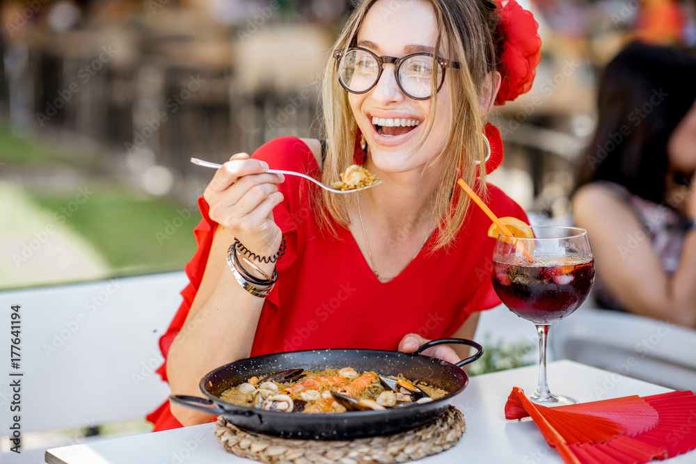 一位身穿红色连衣裙的年轻女士坐在户外吃着传统的巴伦西亚米饭——海鲜饭