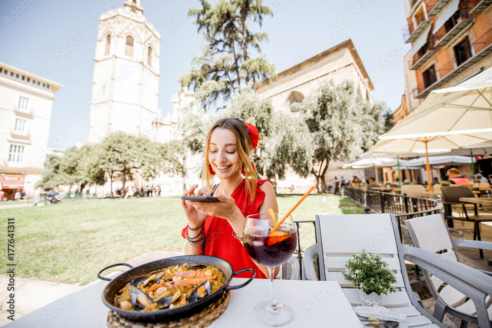 身穿红色连衣裙的年轻女子坐在户外拍摄传统的巴伦西亚米饭——海鲜饭