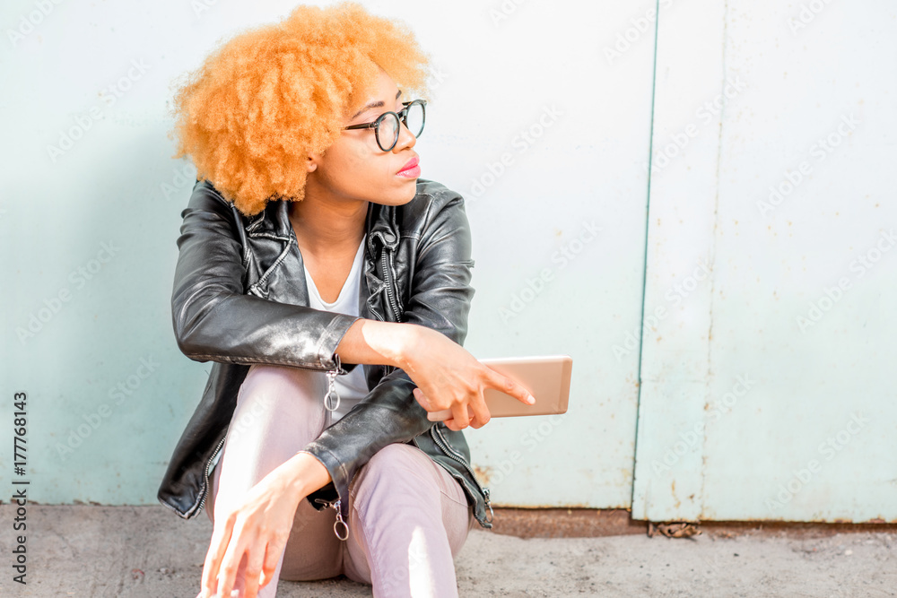 一位身穿皮夹克的非洲妇女的生活方式画像，她坐在绿色的墙上，手里拿着手机。