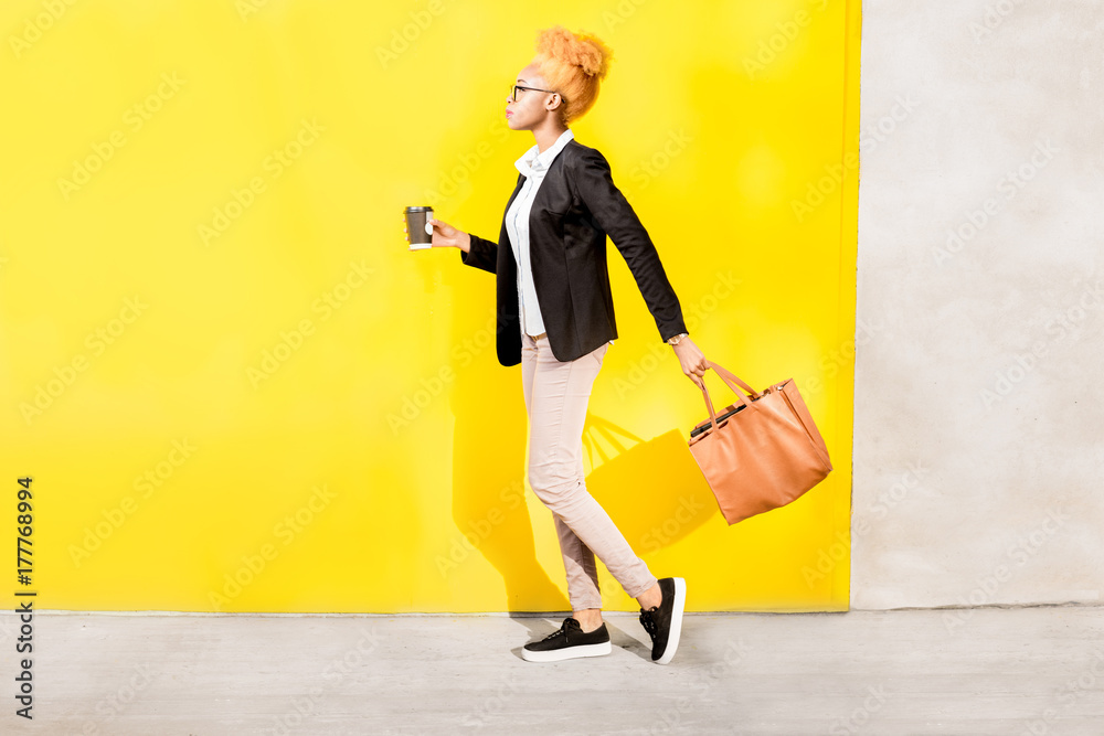 一位穿着休闲西装、提着包走在黄色墙上的非洲商务女士的全身照