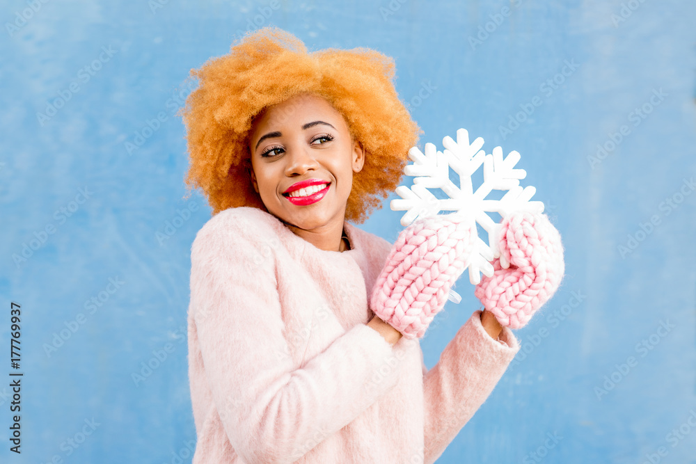 Portrait of a cute african woman in the pink coat holding an artificial snowflake on the blue wall b