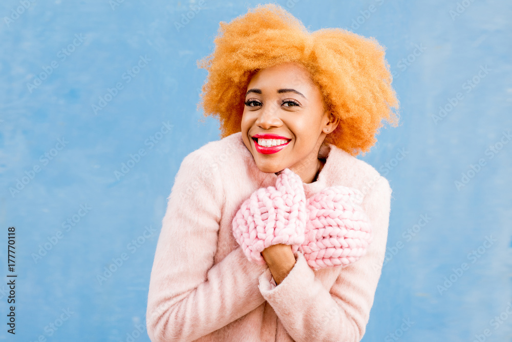 蓝色墙壁背景上一位身穿粉色外套、戴着针织手套的可爱非洲女性的肖像。Wi