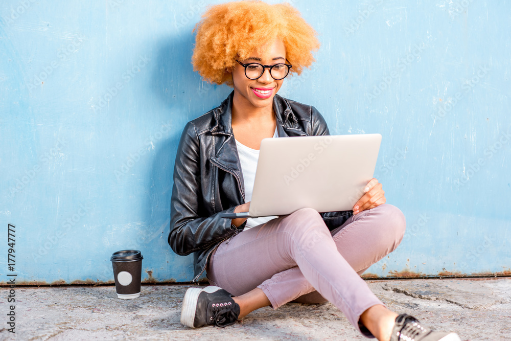 一位留着卷发的非洲妇女坐在蓝色墙壁背景上拿着笔记本电脑工作