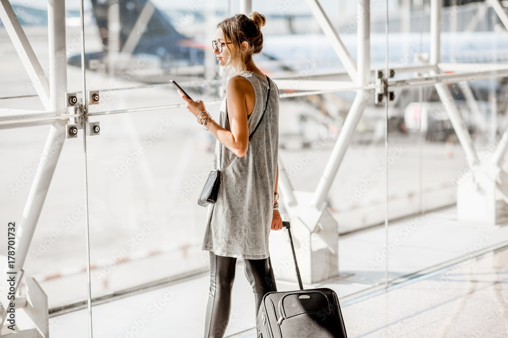 一名年轻女子拿着手机站在机场窗口附近等待航班