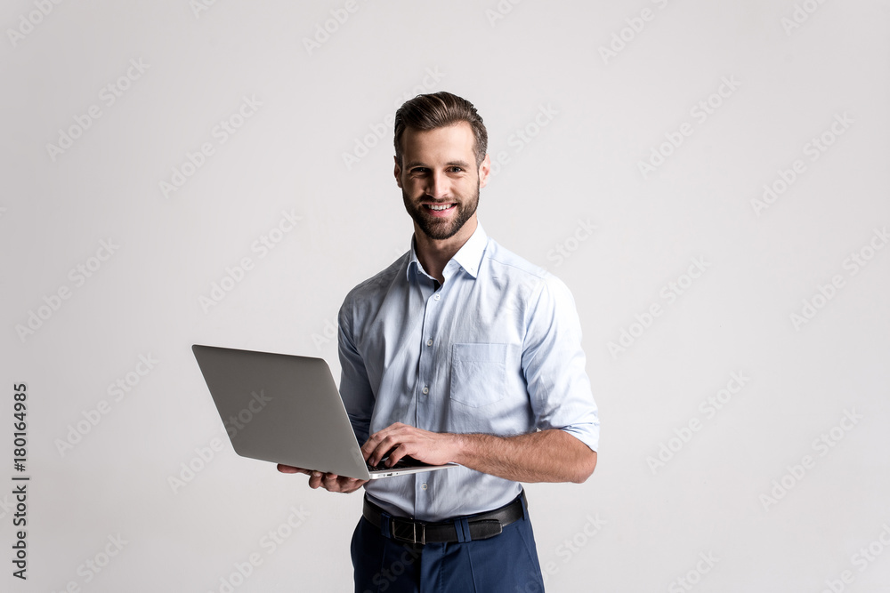 快乐地工作。英俊的年轻人站着用笔记本电脑微笑着看着相机