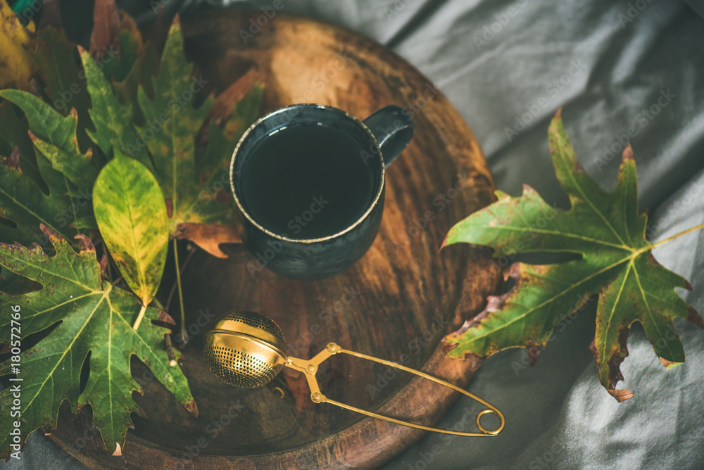 秋天的早茶躺在床上。一杯带筛子的红茶和五颜六色的落叶放在乡村的木地板上