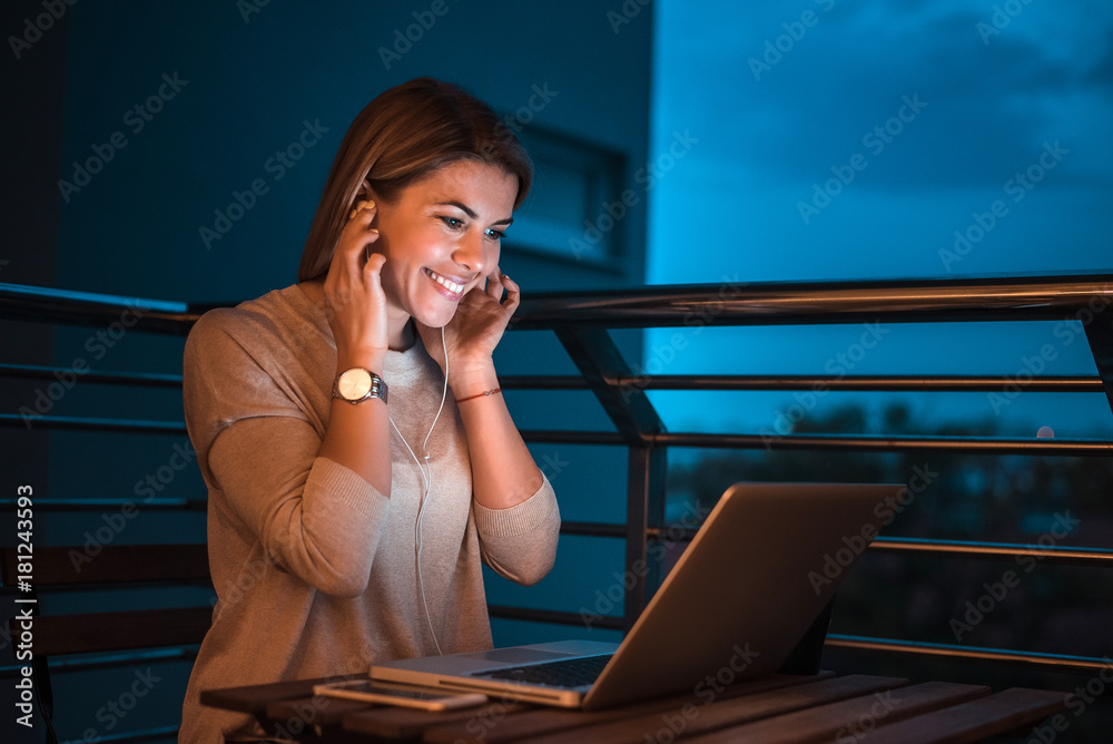 Young blonde woman using laptop late and listening to music. High ISO image.