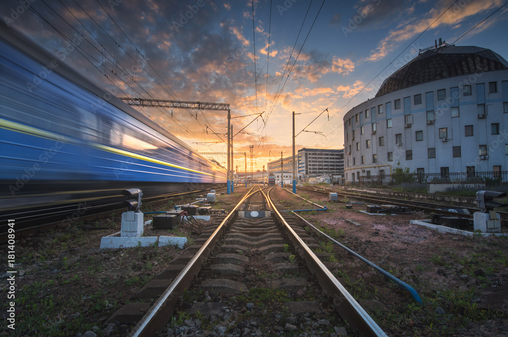 High speed passenger train in motion on railroad track at sunset. Railway station with blurred moder