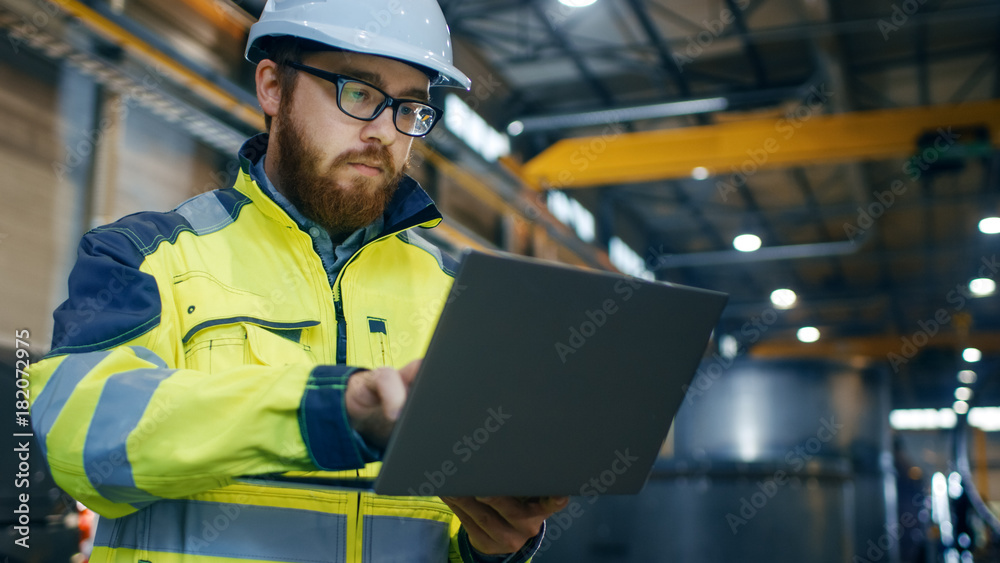 戴着安全帽、穿着安全夹克的工业工程师使用触摸屏笔记本电脑。他在重型工厂工作