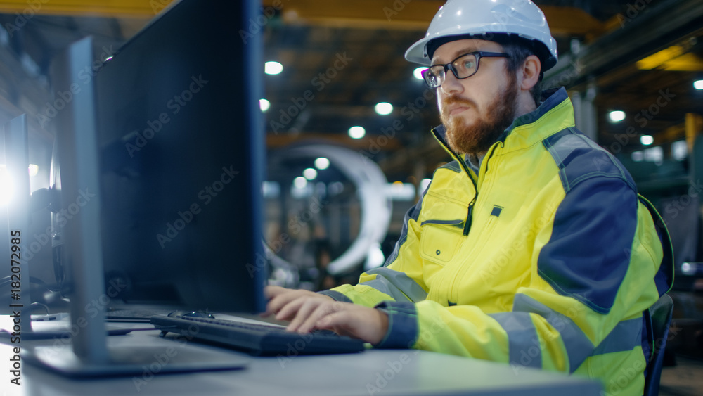 Industrial Engineer Works at Workspace on a Personal Computer.  He Wears Hard Hat and Safety Jacket 