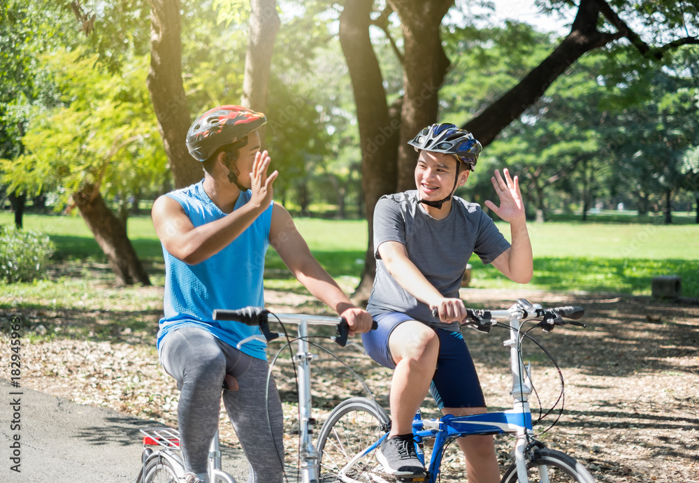 亚洲男孩和朋友夏天在户外绿色公园骑自行车/自行车，互相问好/再见