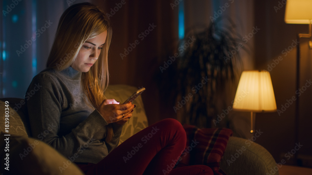 漂亮的年轻女人坐在客厅的沙发上。她饶有兴趣地使用智能手机。它