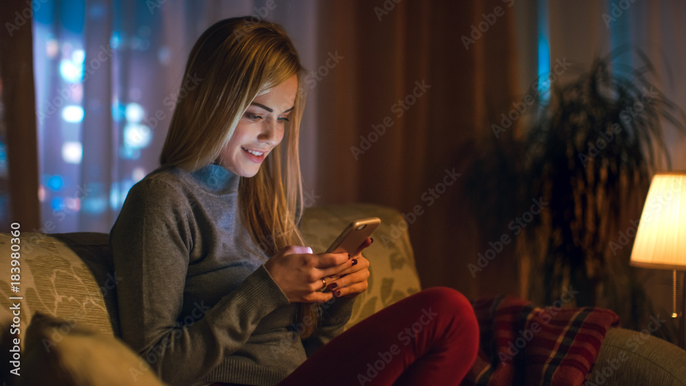 漂亮的年轻女人坐在客厅的沙发上。她饶有兴趣地使用智能手机。它
