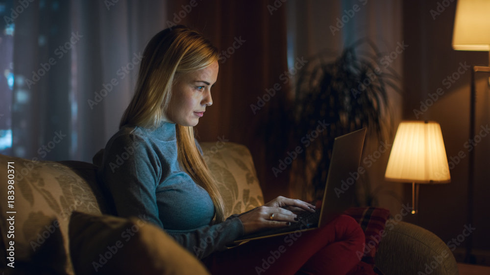 Beautiful Young Woman Works on a Laptop in Her Living Room, She is Sitting on a Sofa with Notebook o