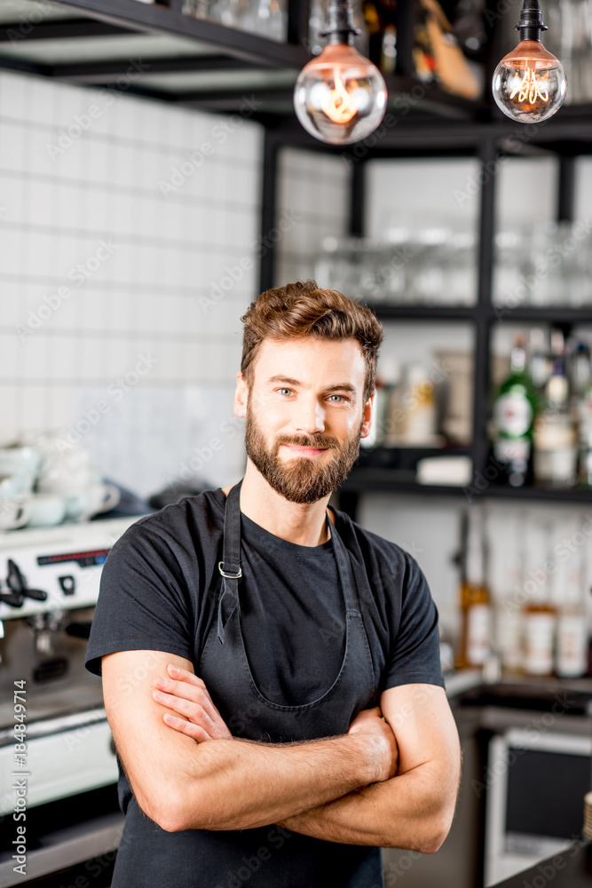 Portrait of a handsome barista in black t-shirt and apron standing at the bar of the modern cafe