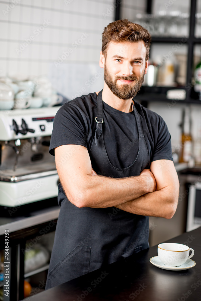 一位穿着黑色t恤和围裙的英俊咖啡师站在现代咖啡馆酒吧的肖像