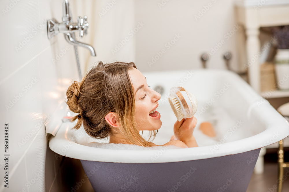 年轻女子在浴室里充满泡沫的漂亮复古浴缸里用刷子唱歌放松