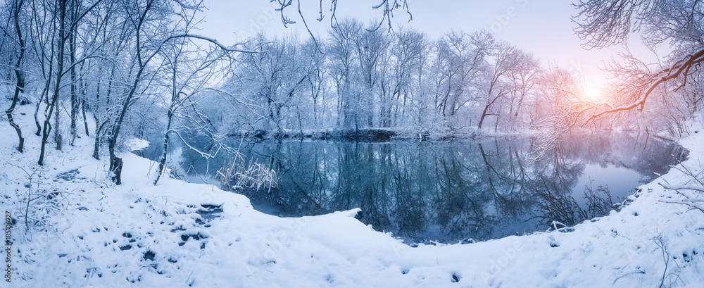 日落时河边的冬季森林。白雪皑皑的树木点缀着五颜六色的景观，河流倒影着