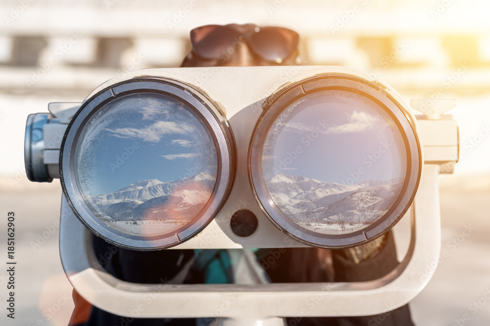 一个女人用双筒望远镜看。在玻璃的反光中，你可以看到雪和山。苏