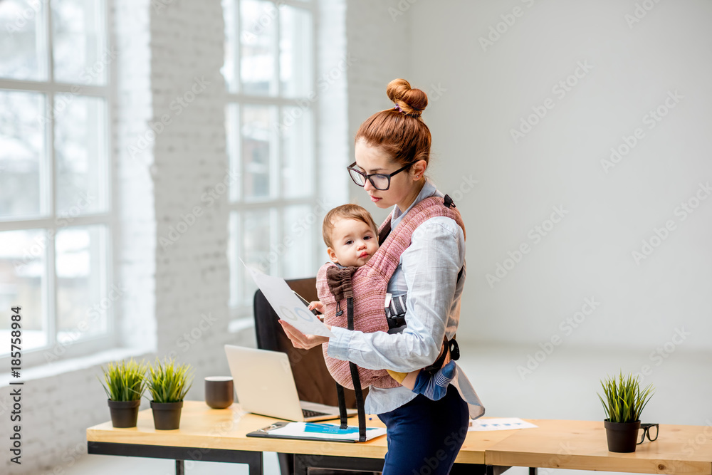 年轻的多任务女商人在办公室工作时带着她的小儿子站着