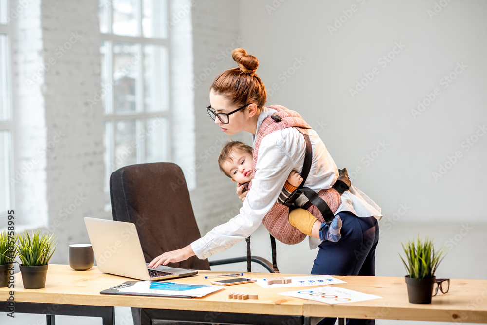 Young multitasking businesswoman standing with her baby son during the work at the office
