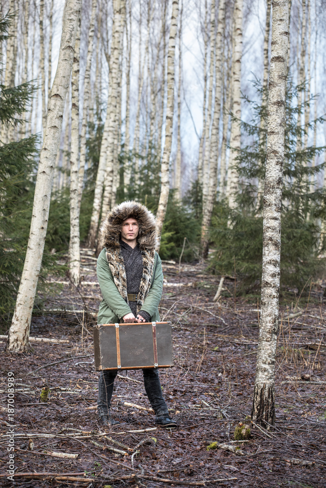 一个年轻人在森林里穿着绿色的冬季派克大衣，提着行李箱。