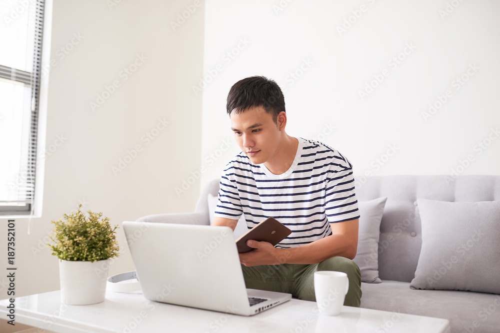 英俊严肃的男人，看着笔记本电脑屏幕，在笔记本上写东西，坐在沙发上