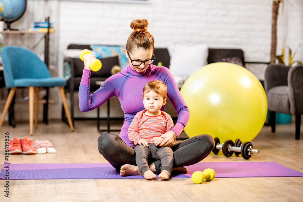 Young mother in sportswear doing exercise lifting a dumbbell with her baby son on the mat at home