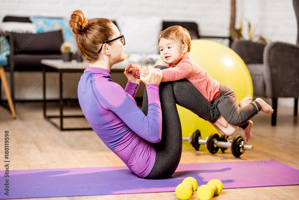 Young mother in sportswear doing exercise lifting with legs her baby son on the mat at home