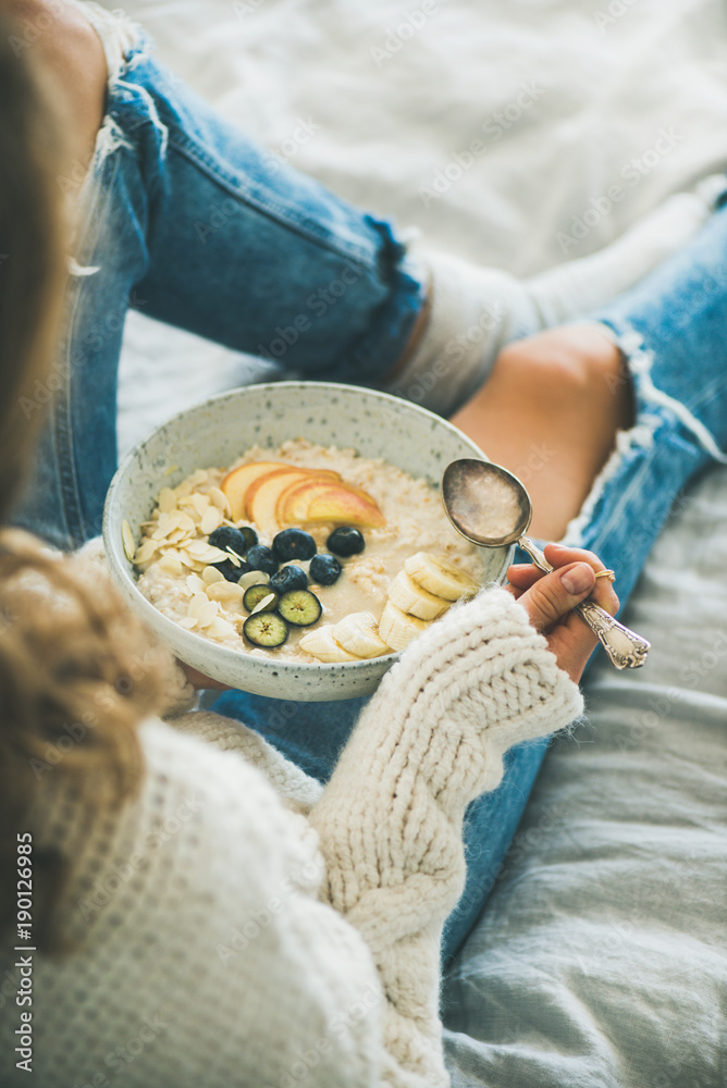 床上的健康冬季早餐。穿着羊毛衫和破旧牛仔裤的女人在吃纯素食杏仁奶o