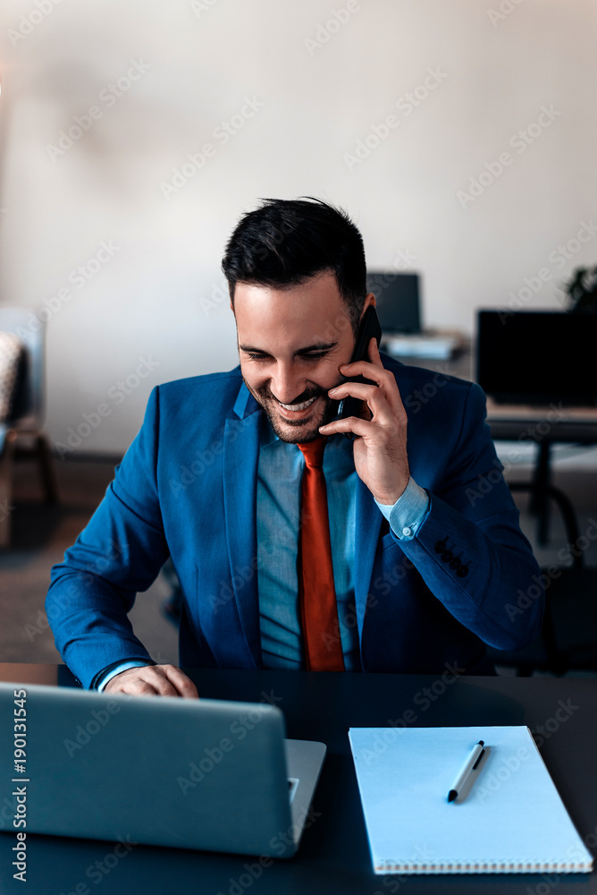 有魅力的白人商人拿着笔记本电脑坐在办公桌前使用手机。