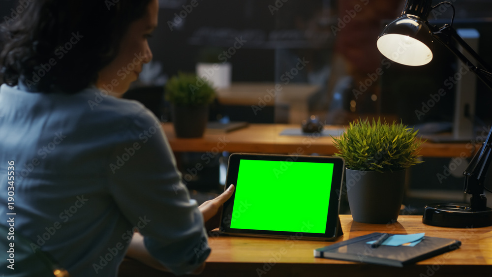 年轻女子坐在她的木制桌子旁，使用带有绿色屏幕的平板电脑，她使用各种各样的