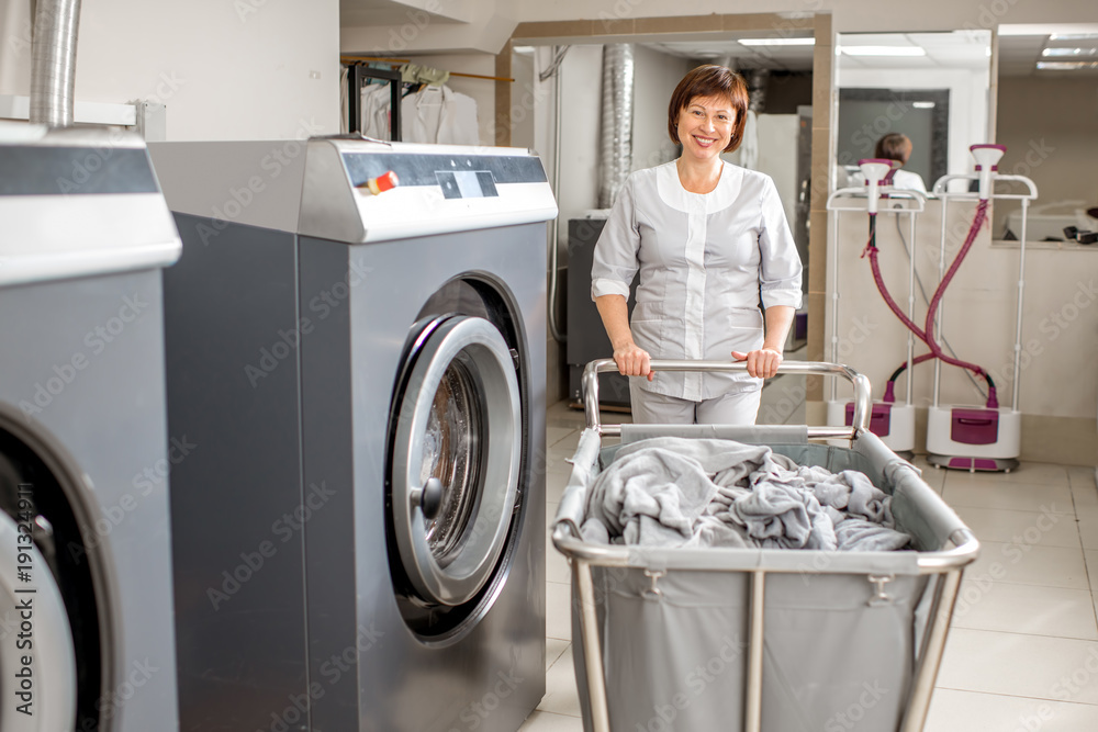 Senior washwoman in uniform standing with clothes basket near the wahing machine in the professional