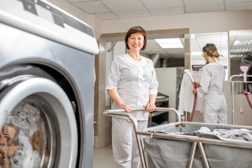 穿着制服的资深洗衣女工拿着洗衣篮站在专业洗衣机附近