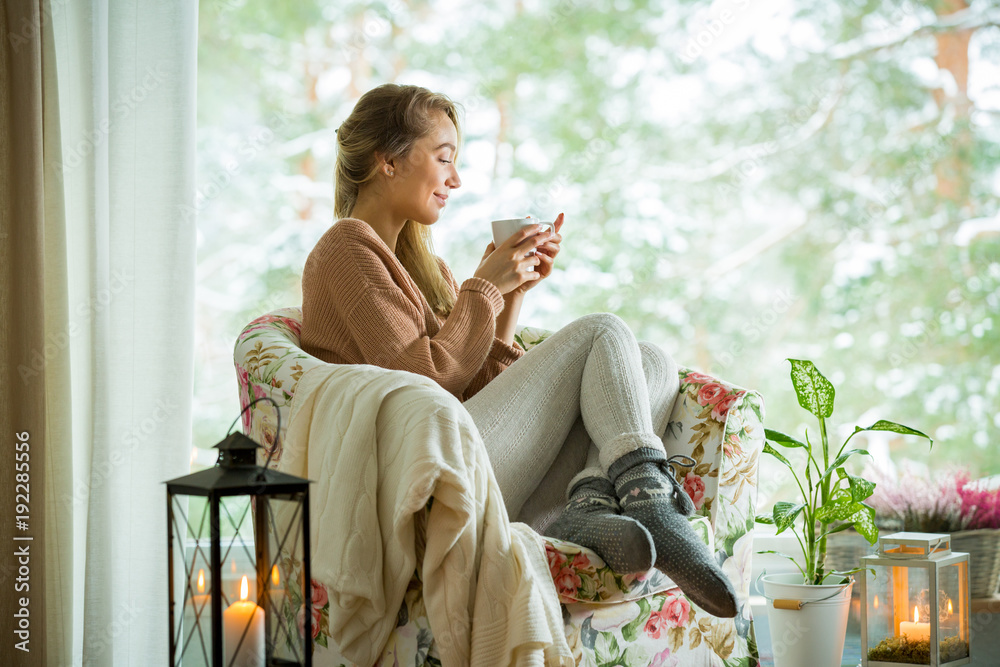 年轻女子穿着针织保暖汗衫，坐在家里靠窗的椅子上，喝着一杯热咖啡